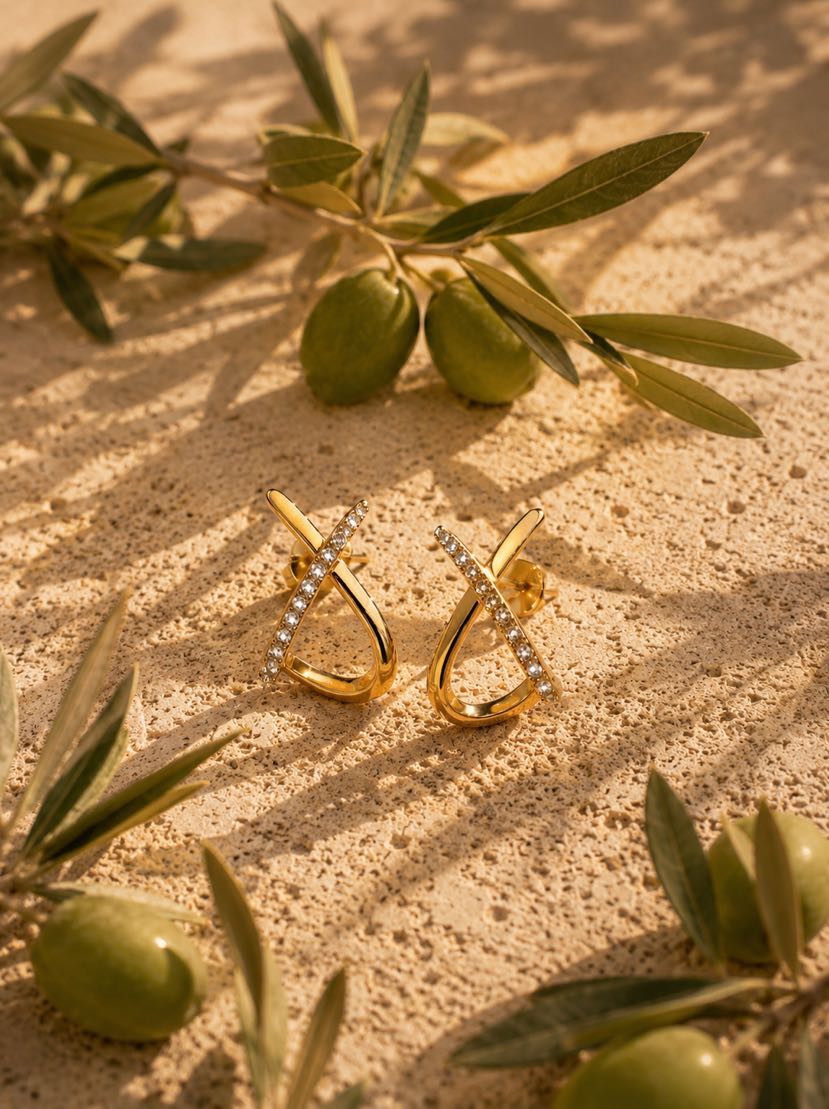 Boucles Divina en X dorées posées sur un bloc de travertin crème, branches d'olivier avec olives vertes en relief autour, ombres dansantes de feuillage, lumière dorée d'après-midi
