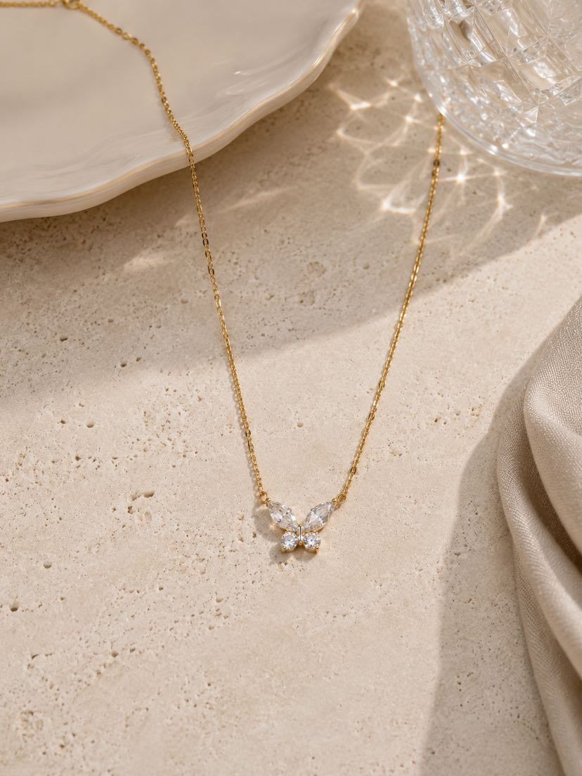 Collier Elyra à plat sur dalle de travertin crème, chaîne fine maillons forçat dorée étirée en arc, pendentif papillon centré au bas serti de quatre topazes blanches taille marquise et perle ronde centrale, assiette en céramique blanche ondulée en haut à gauche et verre en cristal taillé en haut à droite avec reflets prismatiques, drap de lin écru rayonnant à droite