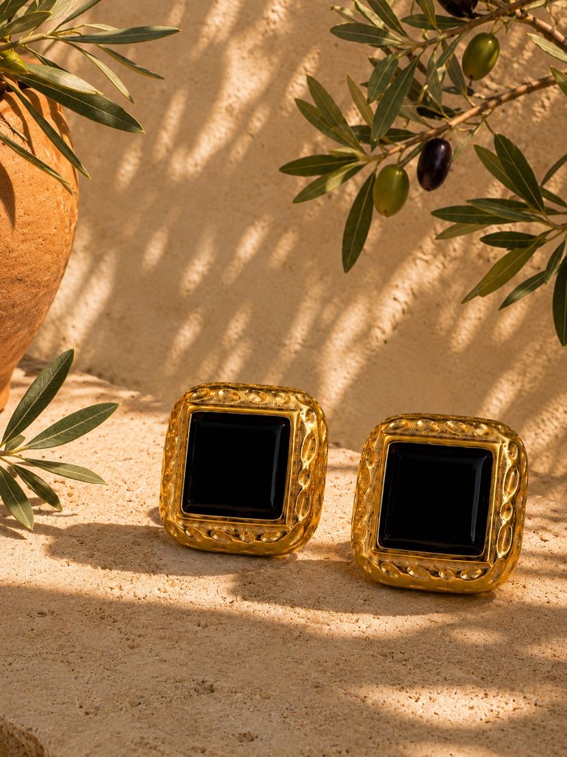 Boucles Paloma carrées en plaqué or à bordure ciselée et cabochon onyx noir, posées sur un bloc de travertin crème au pied d'une jarre en terracotta, branche d'oliviers avec olives vertes et noires en arrière-plan, ombres mouchetées du soleil de fin d'après-midi sur mur ocre