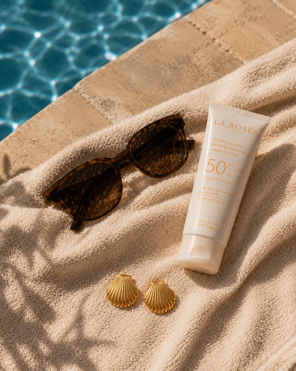 Boucles Solea posées sur une serviette en éponge écrue, lunettes en écaille et tube de crème solaire La Rosée, bord de piscine en travertin