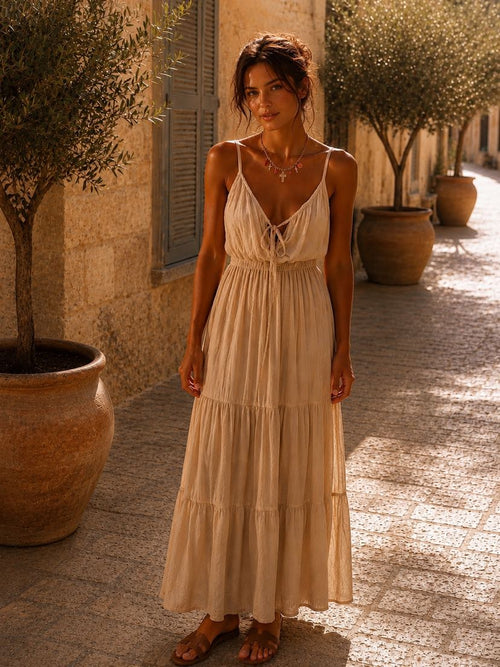 Brune chignon flou et mèches au vent en robe d'été crème en coton et dentelle à fines bretelles avec décolleté en V noué, collier Corallia porté à la clavicule, dans une ruelle pavée méditerranéenne entre oliviers en pots et jarres en terracotta, volets pastel et mur de pierre ocre, lumière dorée d'après-midi