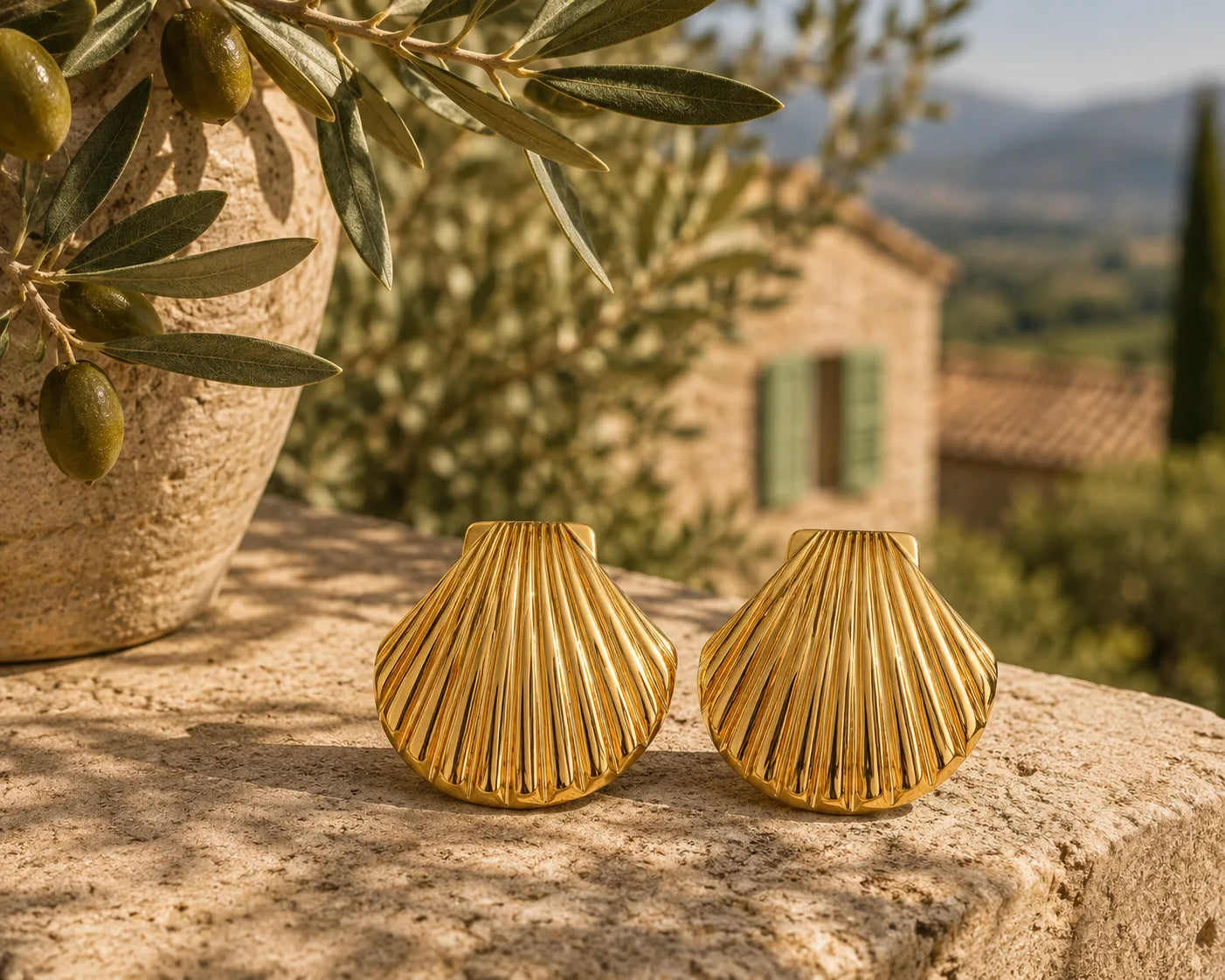 Boucles Solea posées sur un muret de pierre, jarre en terracotta avec branche d'olivier et olives vertes à gauche, bastide provençale aux volets vert pastel et collines en arrière-plan