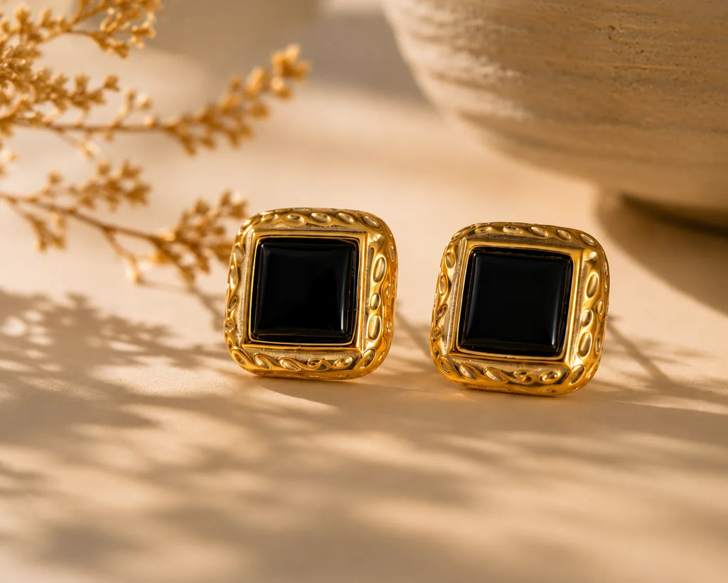 Closeup macro des boucles Paloma carrées en plaqué or à bordure ciselée et cabochon onyx noir, posées sur surface crème, ombre de fleurs séchées, esprit d'atelier