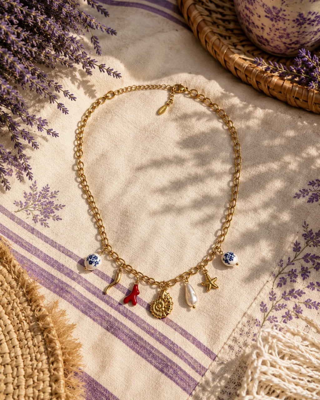 Collier Riviera posé sur un torchon de lin écru rayé de lavande, brins de lavande à gauche, panier en paille et coupe en céramique de Moustiers à droite, ombre de feuillage sur la table