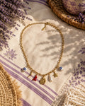 Collier Riviera posé sur un torchon de lin écru rayé de lavande, brins de lavande à gauche, panier en paille et coupe en céramique de Moustiers à droite, ombre de feuillage sur la table