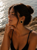 Portrait au bord de la mer, peau dorée, chignon flou, bretelle noire, boucle Paloma carrée or et onyx noir à l'oreille, lumière dorée du couchant sur la mer scintillante